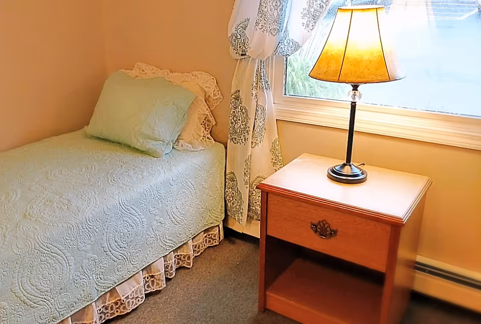 A small bedroom with a single bed covered in a light green quilt and matching pillow. Next to the bed is a wooden nightstand with a drawer and an open shelf, topped with a lit table lamp. A window with patterned curtains is behind the nightstand.