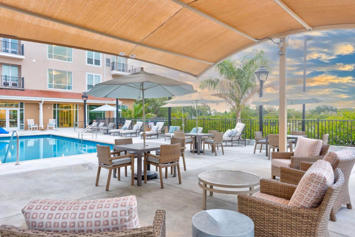Outdoor poolside patio with tables, umbrellas, wicker lounge chairs and a covered seating area beside a residential building.