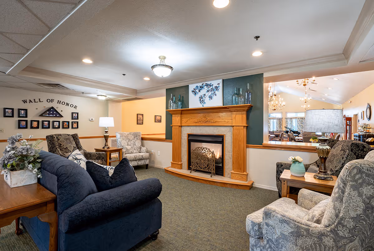 A cozy living room area in a senior living facility featuring a fireplace with a wooden mantel, surrounded by comfortable upholstered chairs and a dark blue sofa. The wall above the fireplace is decorated with art and glass bottles. To the left, there is a 'Wall of Honor' with framed photos and a folded American flag. The room is well-lit with ceiling lights and lamps, and a dining area is visible through a large interior window.