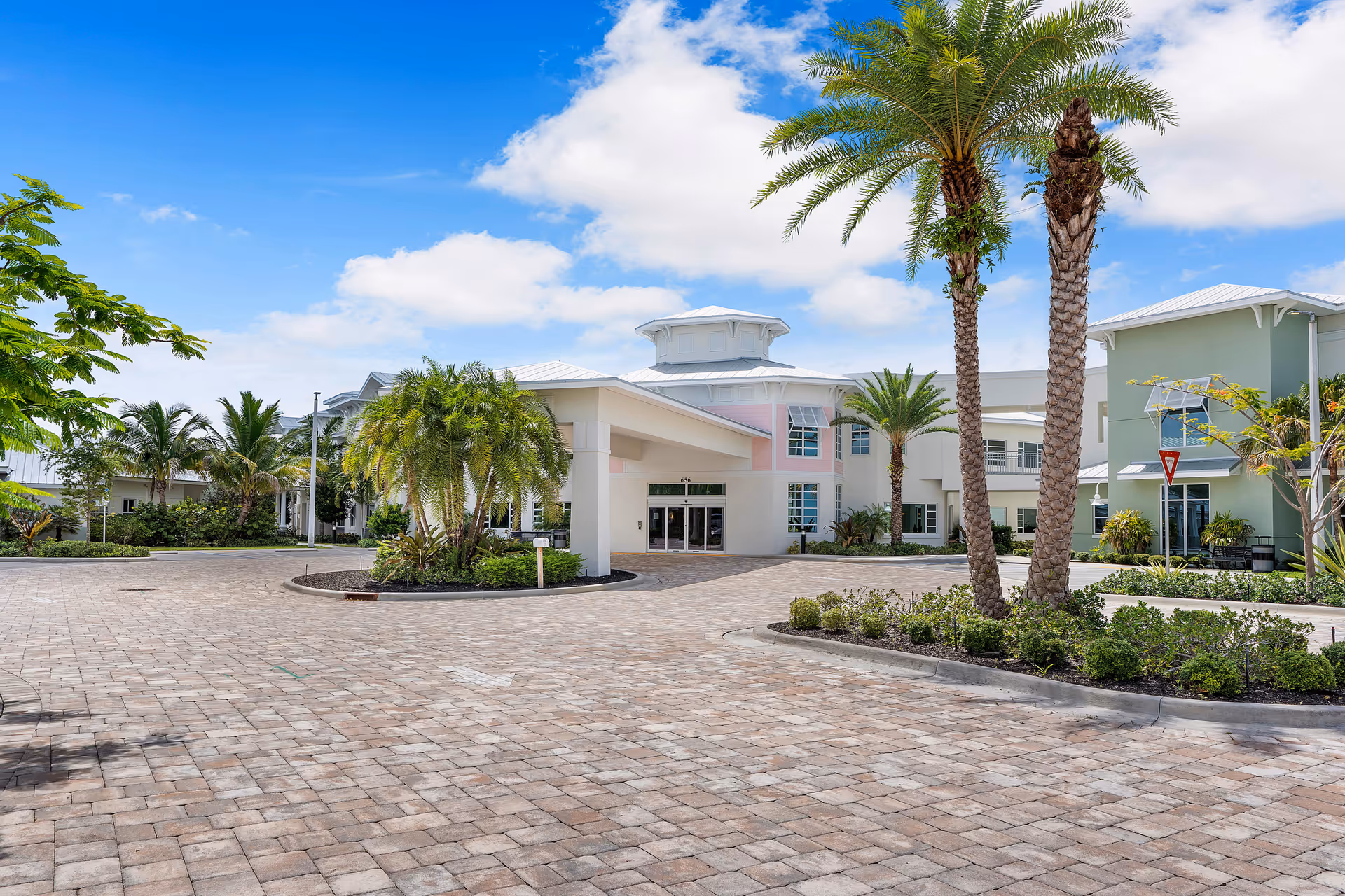 Exterior view of Luxe Senior Living at Jupiter showing a paved driveway with landscaped greenery and palm trees leading to the main entrance of a modern, light-colored building under a blue sky with some clouds.