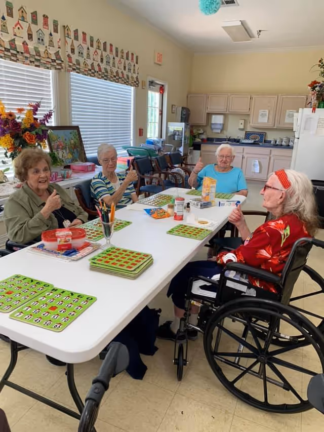 Four elderly women sitting around a white table in a bright room, playing a game with green bingo cards. Three women are seated in regular chairs and one woman is in a wheelchair. They are smiling and giving thumbs up. The room has windows with blinds, a kitchen area in the background, and colorful decorations including flowers and a painting.