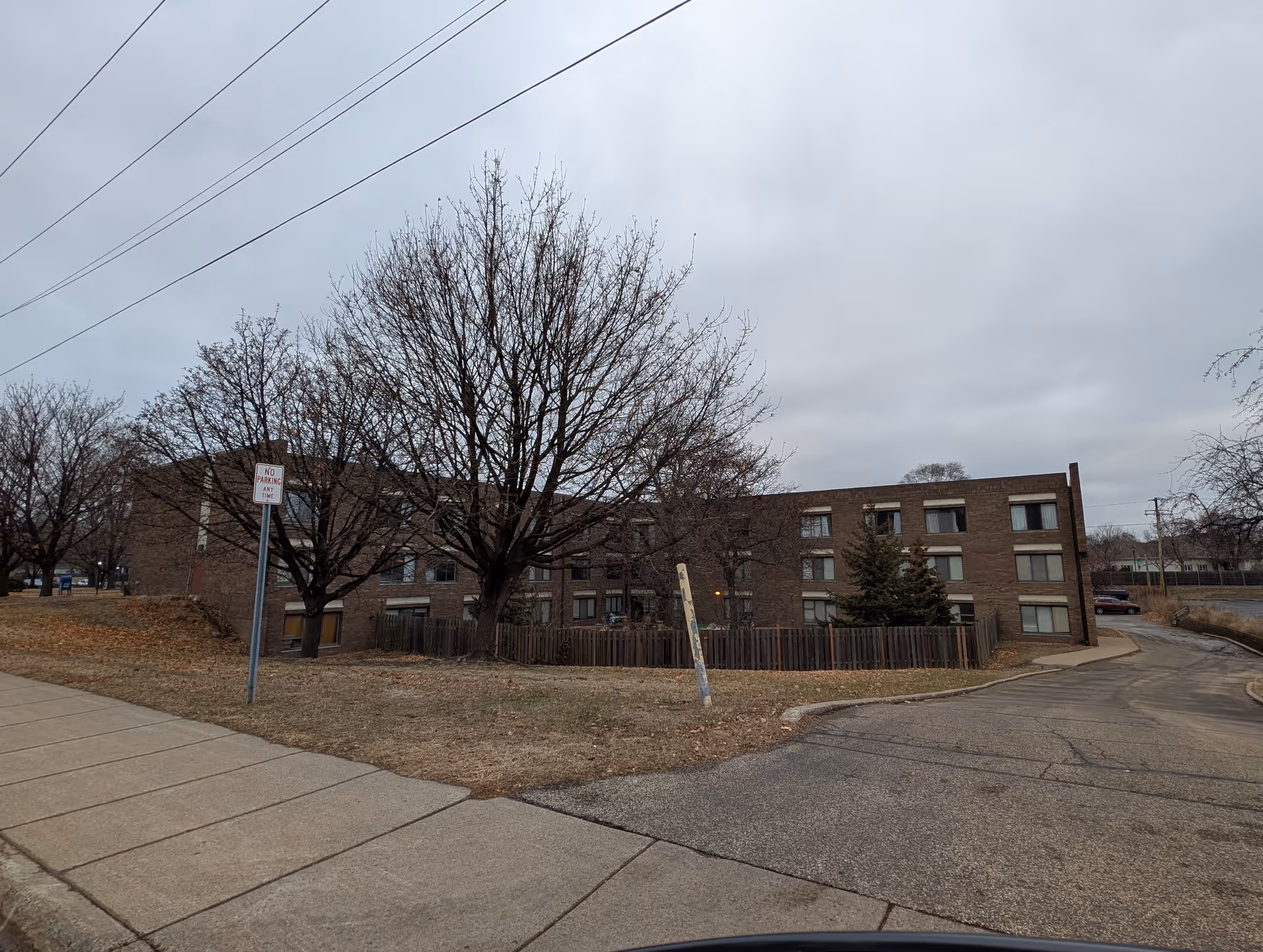 Exterior view of a three-story brick building with multiple windows, surrounded by leafless trees and a fenced area. The sky is overcast and there is a sidewalk and a paved driveway in the foreground.