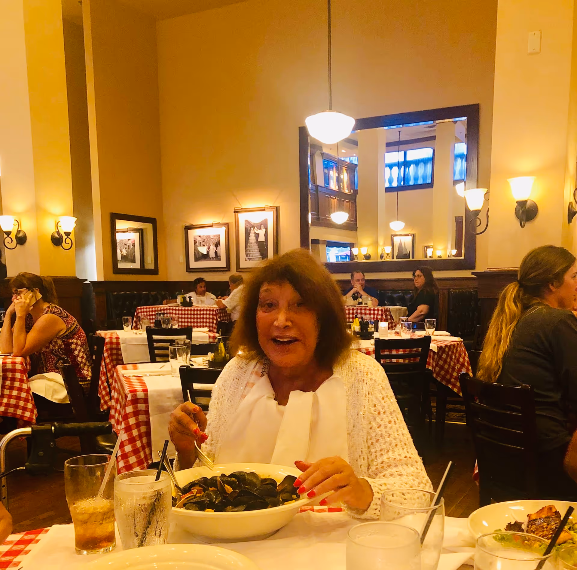 A woman smiling at a table in a busy restaurant with red-and-white checkered tablecloths and a bowl of mussels.