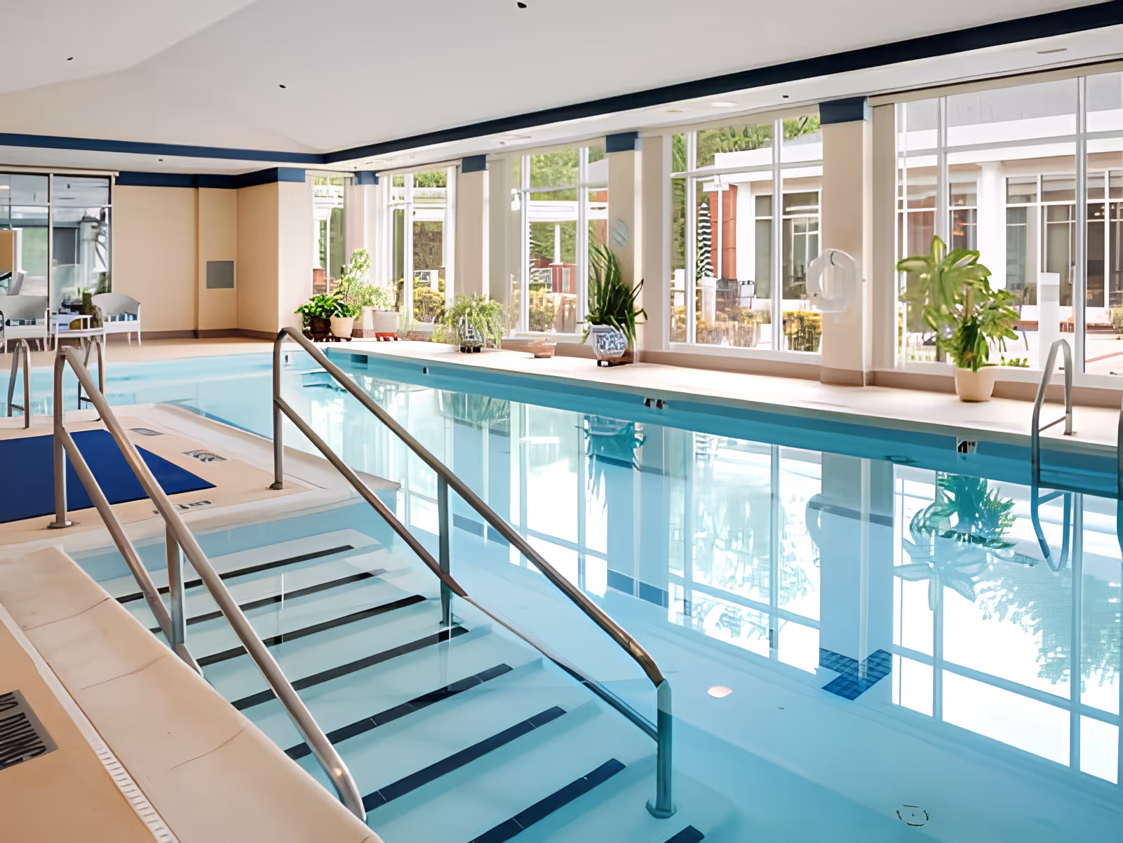 Indoor swimming pool with clear blue water, metal handrails, and steps leading into the pool. Large windows along one side let in natural light and show an outdoor patio area. Several potted plants are placed around the pool area, and there are chairs and tables visible in the background.