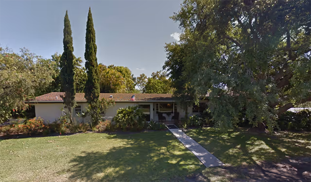 Single-story Sun Bay Manor building with a central walkway, lawn, and mature trees.