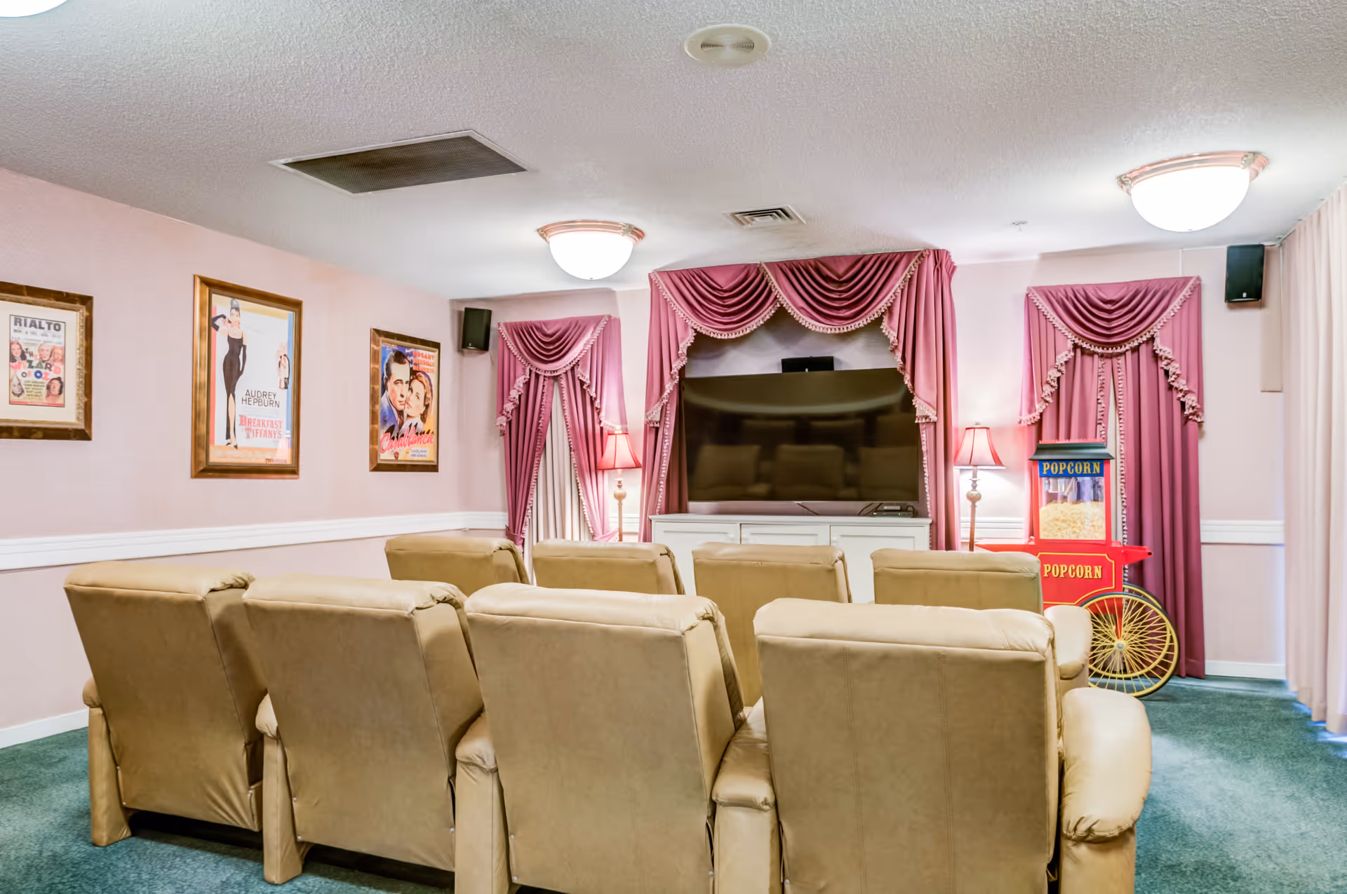 A cozy home theater room with two rows of beige recliner chairs facing a large flat-screen TV. The walls are decorated with framed vintage movie posters, and the windows are adorned with pink drapes with valances. There is a red popcorn machine on the right side of the room, and the carpet is green.