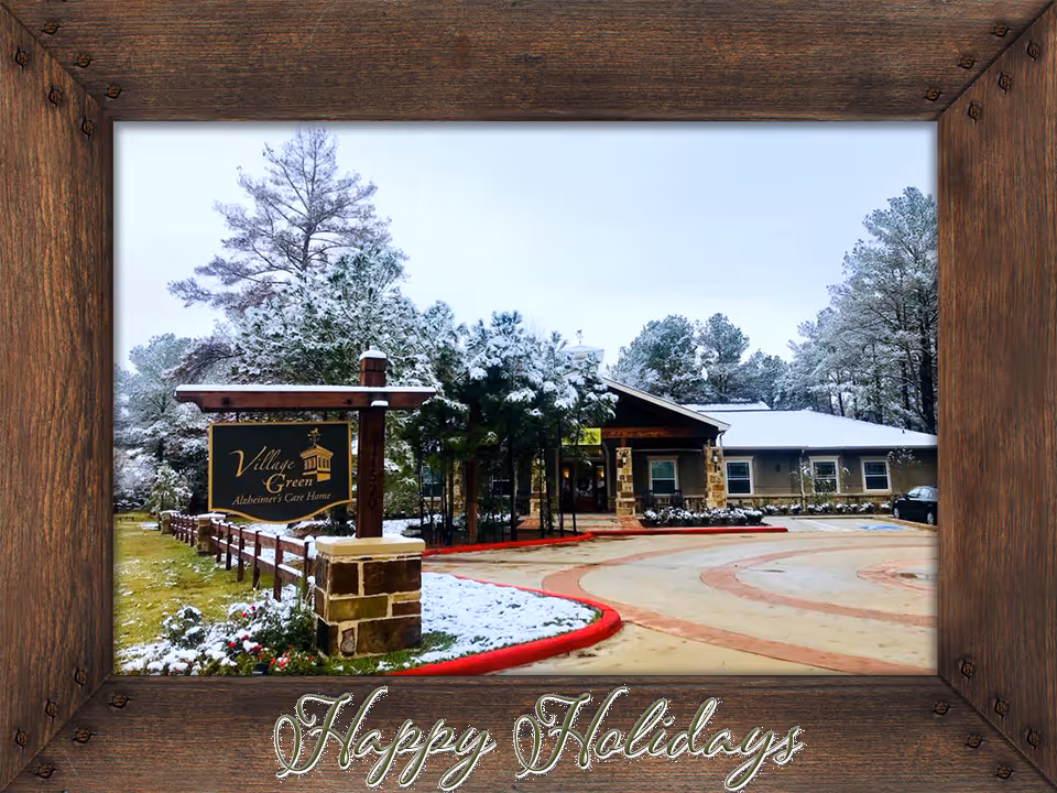 Exterior view of Village Green Alzheimer's Care Home in a snowy setting with trees and a driveway. The building is surrounded by snow-covered ground and greenery. The image is framed with a wooden border and has the text 'Happy Holidays' at the bottom.