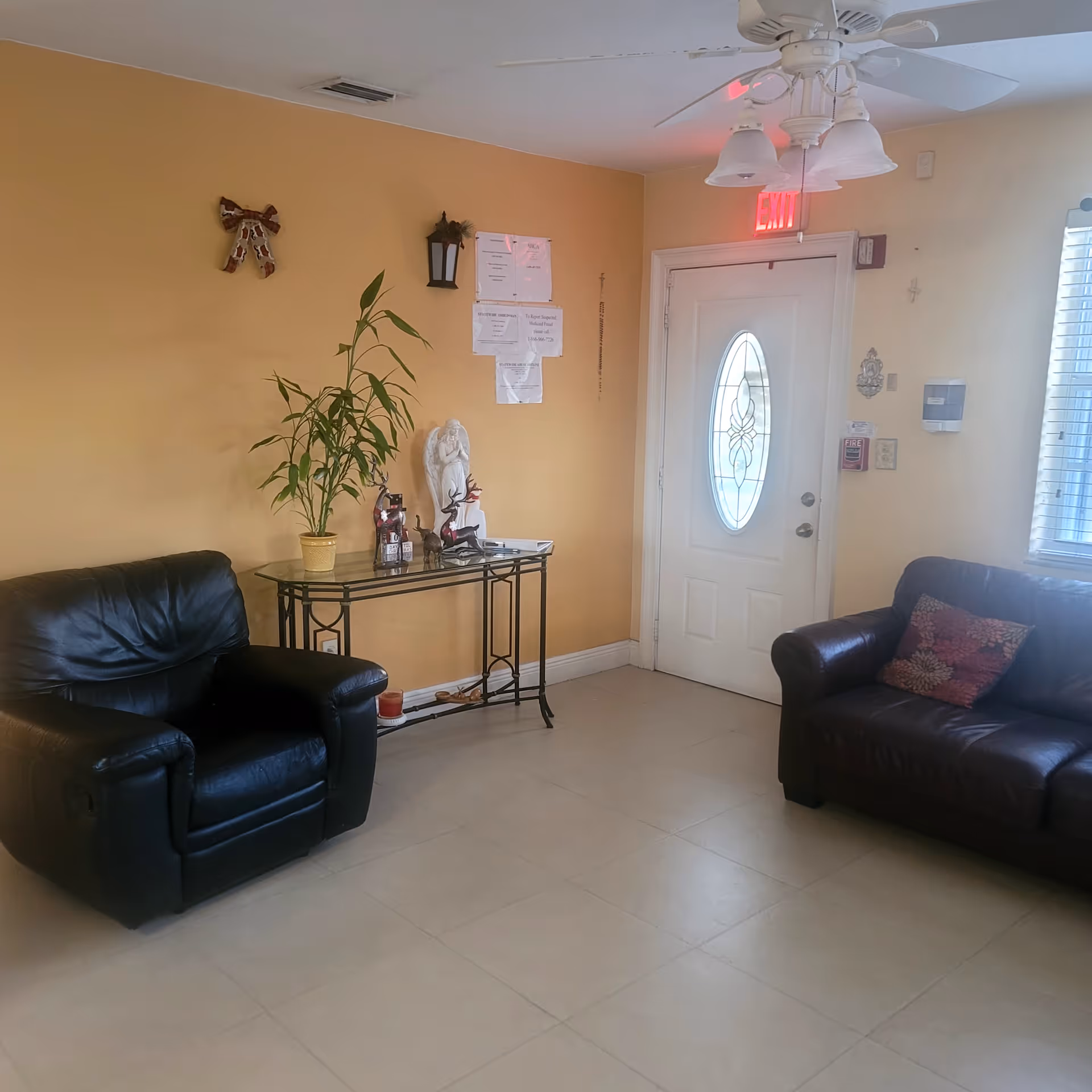 A small living room area with two black leather chairs, a glass table with decorative items and a potted plant, a white door with an oval glass window, and a ceiling fan with lights. The walls are painted yellow and there is an exit sign above the door.