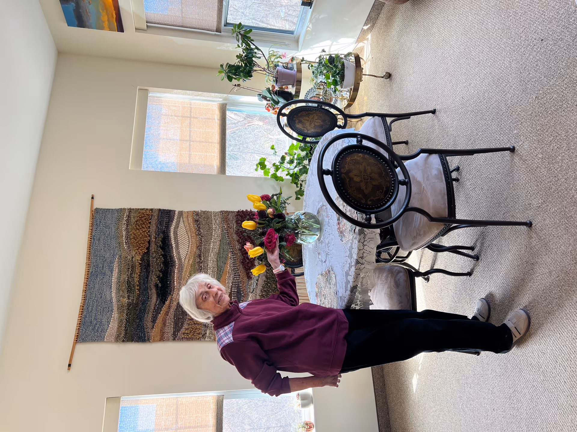 An elderly woman stands beside a round dining table with a lace tablecloth and a vase of tulips in a bright room with plants and a woven wall hanging.