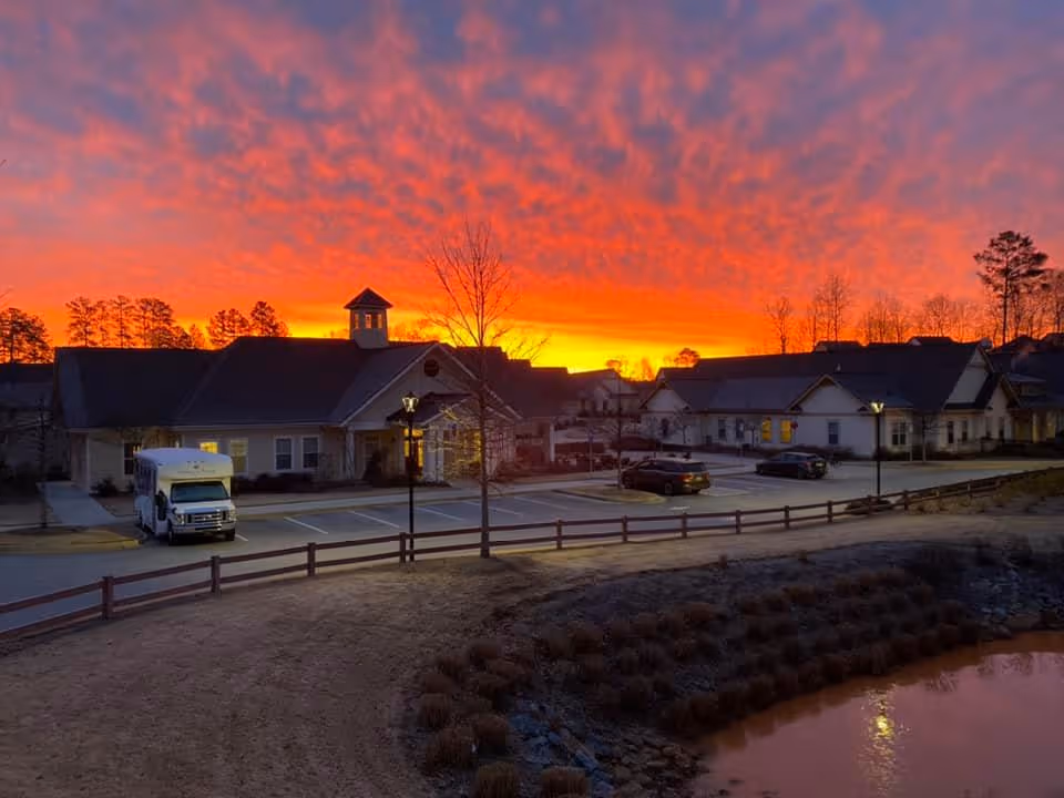 Exterior view of Camellia Place an Oaks Senior Living Community at sunset with a vibrant orange and purple sky. The image shows multiple buildings, a parking lot with cars and a shuttle bus, street lamps, and a small pond in the foreground.