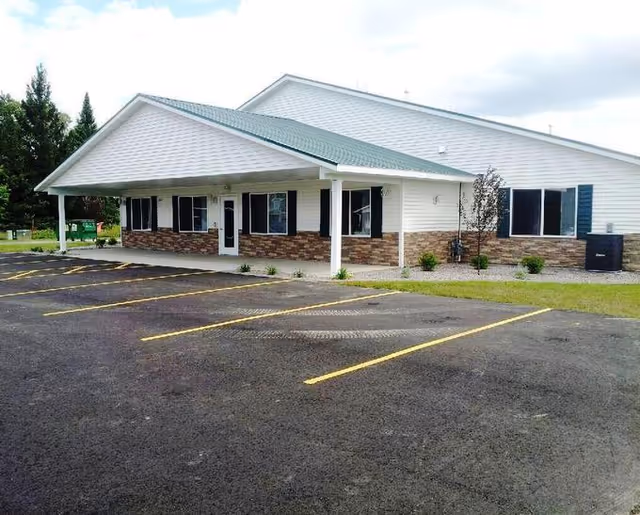 Single-story building with white siding and a green roof, featuring a covered entrance supported by white columns. The building has several windows with dark shutters and a brick facade along the lower half. In front of the building is an empty parking lot with yellow parking lines. Some small shrubs and trees are planted near the building.