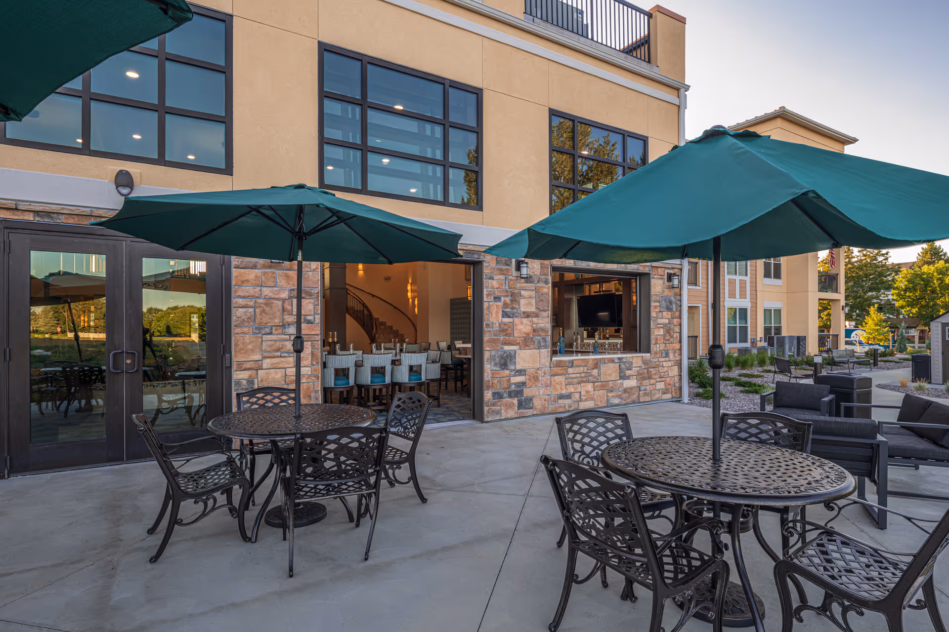 Outdoor patio area at AltaVita Independent Living featuring round metal tables with green umbrellas and matching chairs. The patio is adjacent to a building with large windows and stone accents. Inside, a dining area with chairs and a bar is visible through open doors and a serving window.