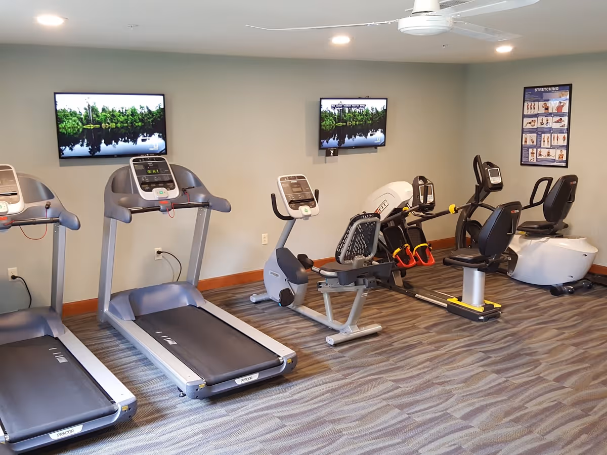 Interior view of a fitness room with two treadmills, two recumbent exercise bikes, and a rowing machine. Two wall-mounted TVs display a nature scene, and a stretching exercise poster is visible on the wall. The room has carpeted flooring and neutral-colored walls.