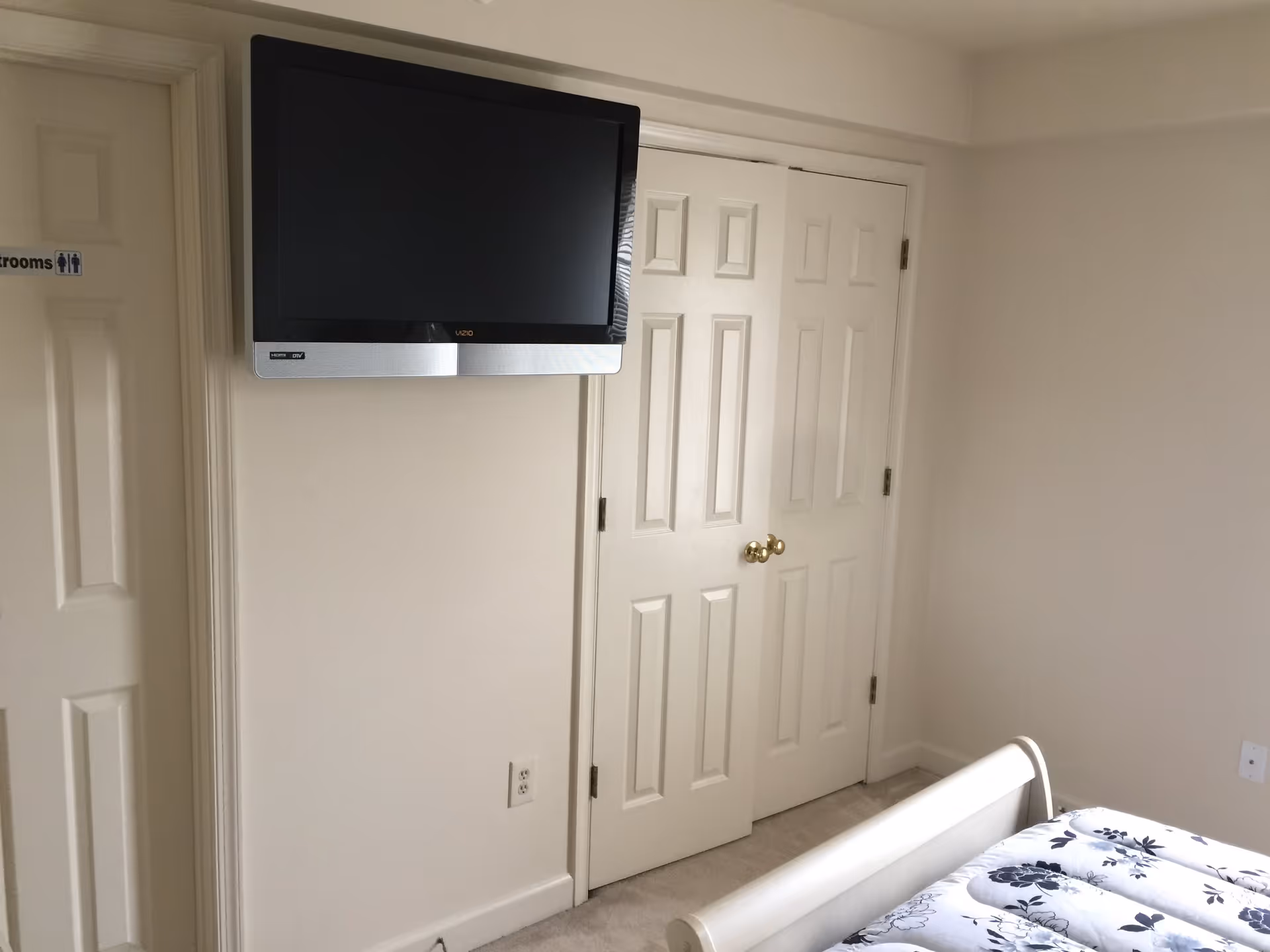 A corner of a bedroom with a mounted flat-screen TV on the wall, a closed double-door closet with gold knobs, part of a bed with a floral-patterned comforter, and a door labeled 'Restrooms'. The walls and doors are painted white.