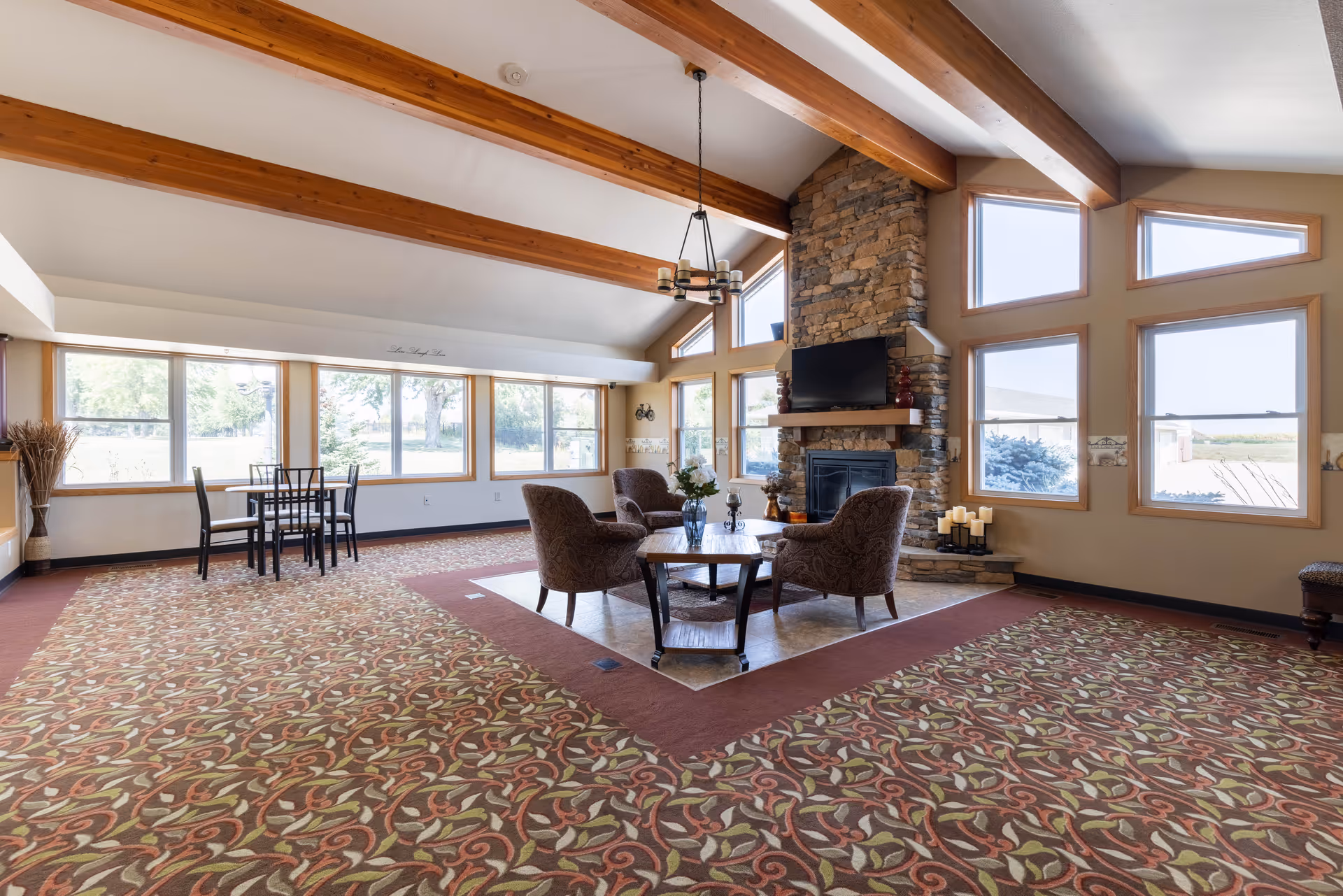 A spacious senior living common area with large windows letting in natural light. The room features a stone fireplace with a mounted TV above it, surrounded by four upholstered armchairs arranged around a wooden coffee table. The ceiling has exposed wooden beams and a chandelier hanging in the center. There is a small dining table with four chairs near the windows, and the floor is covered with a patterned carpet.