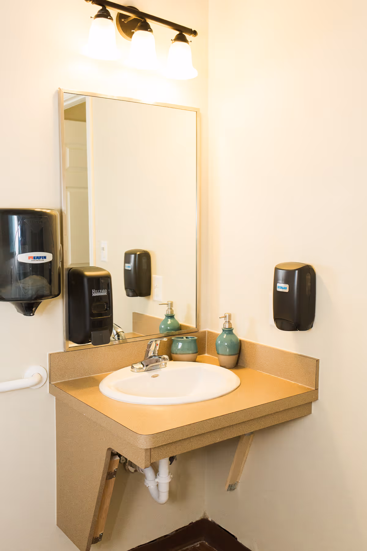 Corner accessible bathroom sink with mirror, wall-mounted soap dispensers, and overhead light.