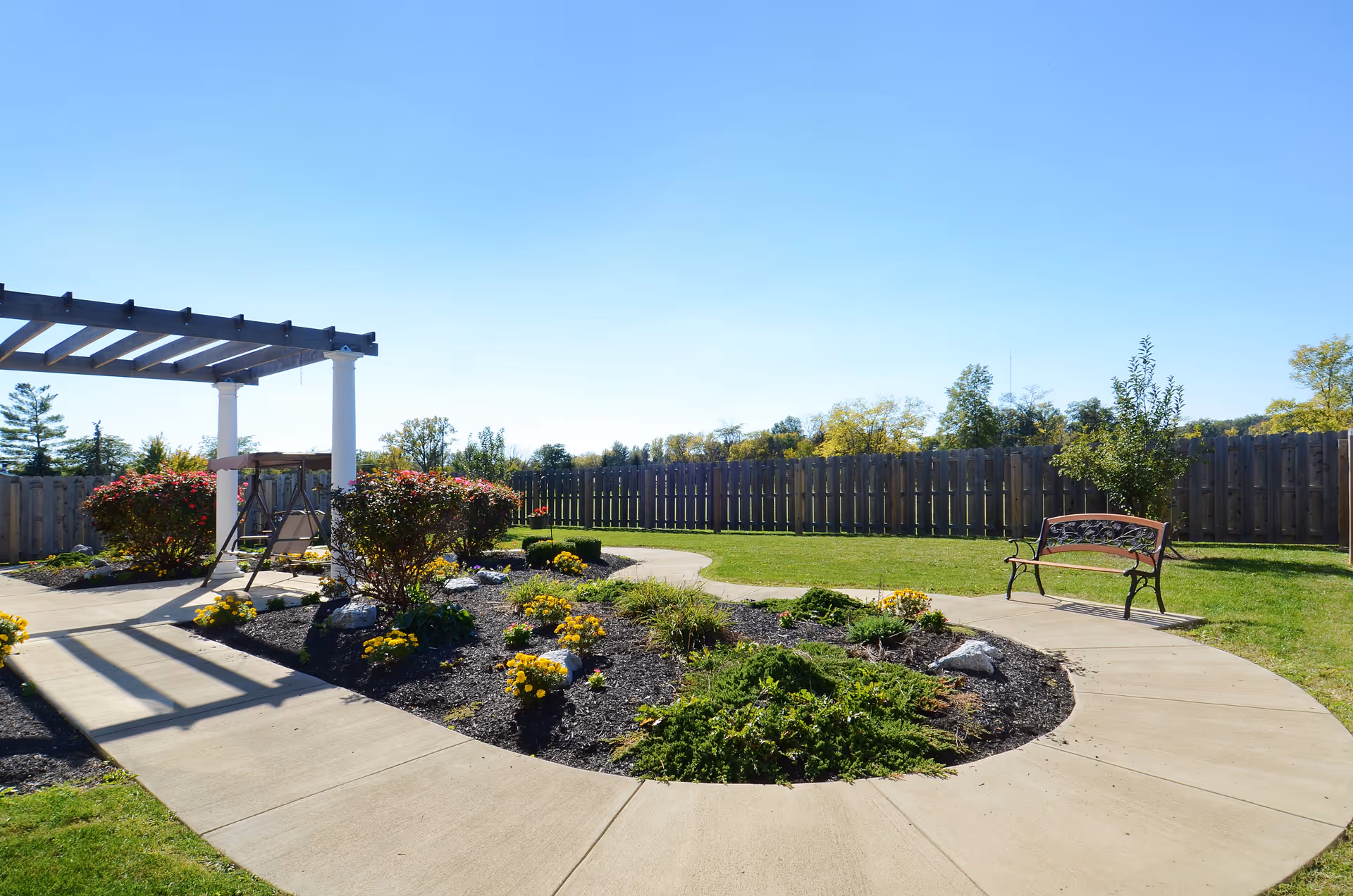 Outdoor garden area with a curved concrete pathway surrounding a landscaped flower bed with bushes and yellow flowers. There is a wooden pergola with a swing underneath it on the left side and a metal bench on the right side. The area is enclosed by a wooden fence with trees visible in the background under a clear blue sky.