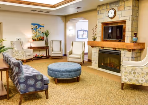A cozy living room area with patterned armchairs and a round blue ottoman in the center. There is a stone fireplace with a wooden mantel, a clock above it, and a TV mounted inside the fireplace structure. The room has warm lighting, a carpeted floor, and decorative plants and artwork on the walls.