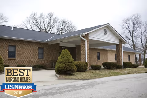 Exterior view of Morris Nursing Home, a single-story brick building with a covered entrance and several windows. There are trimmed bushes and trees around the building, and the sky is overcast.