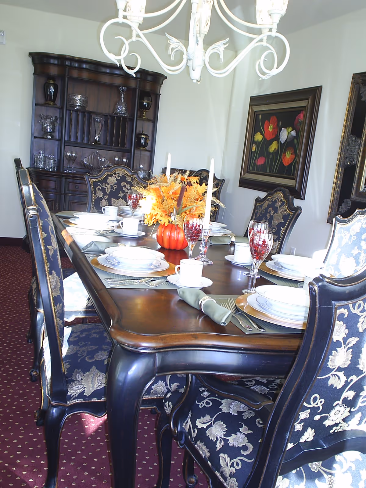 A formal dining room with a long wooden table set for a meal. The table has plates, cups, silverware, wine glasses with red liquid, and a centerpiece featuring a small pumpkin and autumn leaves. The chairs around the table have dark wood frames and patterned upholstery. In the background, there is a dark wooden cabinet with glassware and decorative items. A chandelier hangs above the table, and framed floral artwork is on the wall.