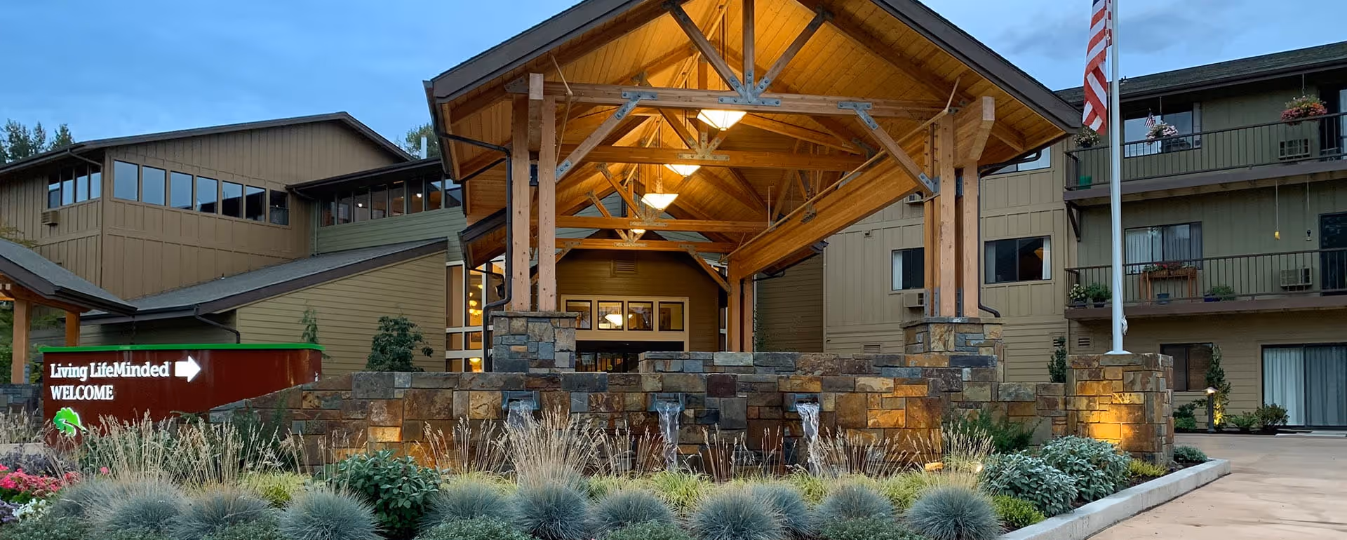 Exterior view of the entrance to Willamette Oaks senior living facility featuring a large wooden canopy supported by stone pillars, a stone water fountain in front, landscaped plants, and an American flag on a flagpole to the right. A sign on the left reads 'Living Life Minded WELCOME' with an arrow pointing to the entrance.