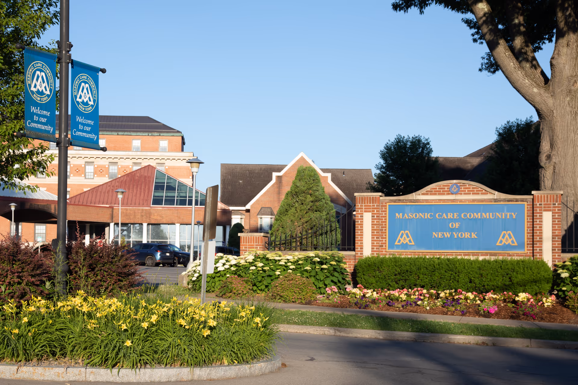 Exterior view of the Masonic Care Community of New York facility with a brick sign displaying the community's name surrounded by well-maintained landscaping including flowers and bushes. The building is visible in the background under a clear blue sky, with banners on lamp posts that say 'Welcome to our Community.'