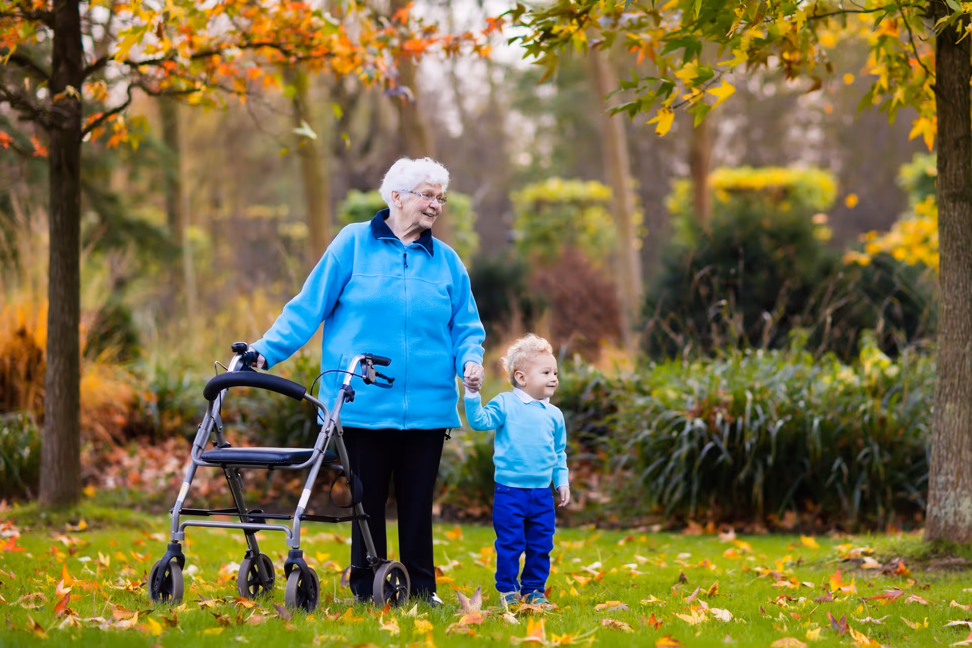 An elderly woman with white hair wearing a blue jacket is standing outdoors in a park-like setting with autumn leaves on the ground. She is holding the hand of a young child dressed in a light blue sweater and dark blue pants. The woman has a walker beside her and both are smiling, surrounded by trees and greenery.