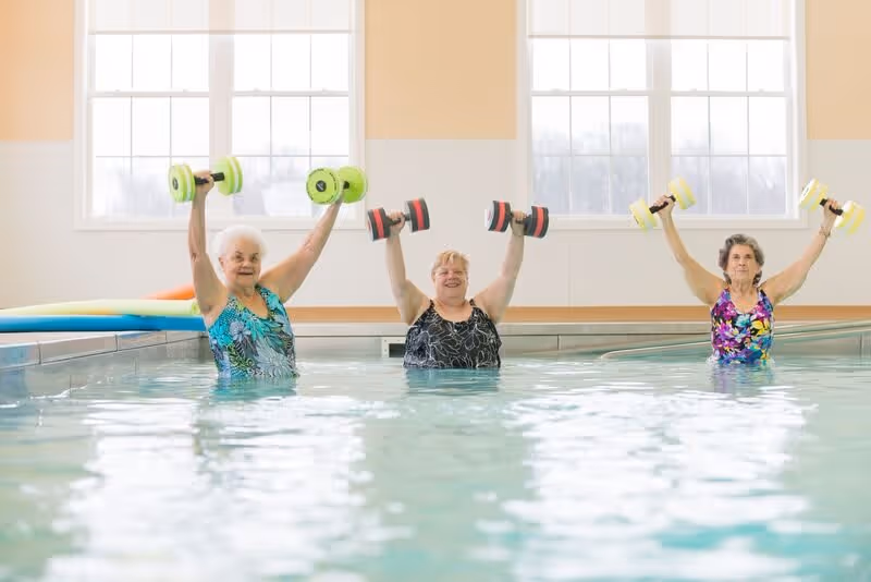 Three elderly women standing in an indoor swimming pool, each holding a pair of colorful water dumbbells raised above their heads, with large windows in the background letting in natural light.