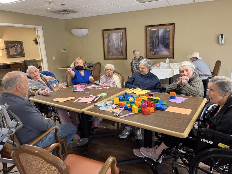 A group of elderly individuals sitting around a large table in a common room, engaging in arts and crafts activities with colorful paper, scissors, and building blocks. The room has beige walls with framed pictures and a few other people seated at a table in the background.