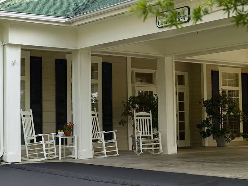 Covered front porch with white rocking chairs, a small table with potted plants, and supporting columns at a building entrance.