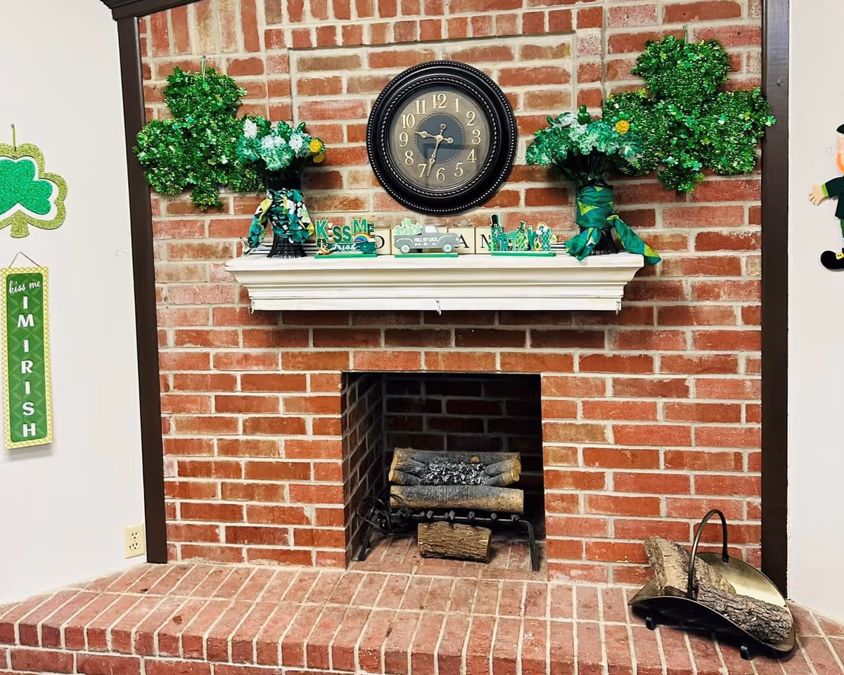 A brick fireplace with a white mantel decorated with St. Patrick's Day themed items including two green shamrock-shaped decorations, two bouquets of white flowers wrapped in green cloth, a clock above the mantel, and various small green decorative pieces. To the left of the fireplace, a green sign reads 'kiss me I M I R I S H'. Firewood and fireplace tools are placed on the hearth.