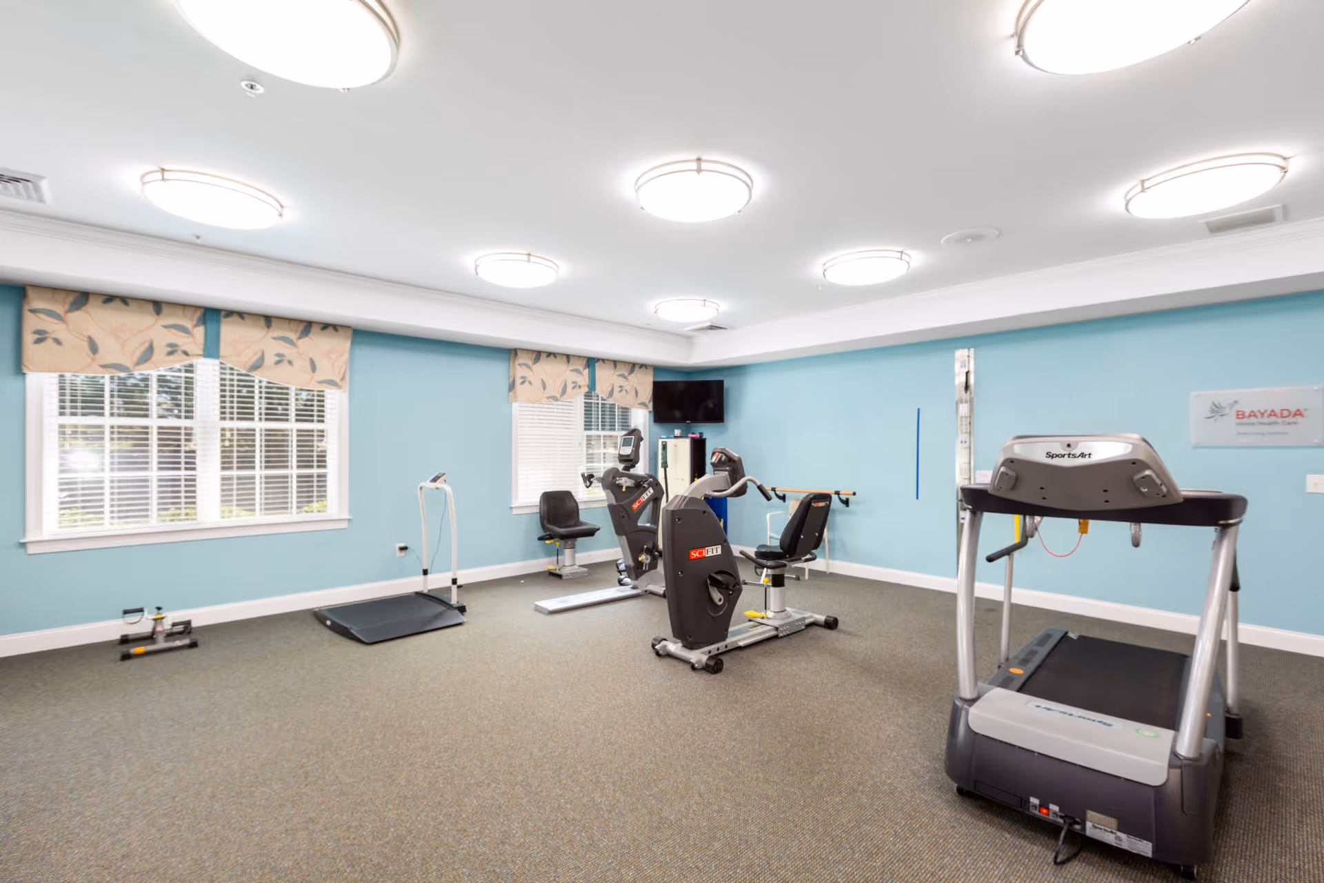 Bright fitness room with a treadmill, recumbent exercise bikes, exercise mat and large windows.
