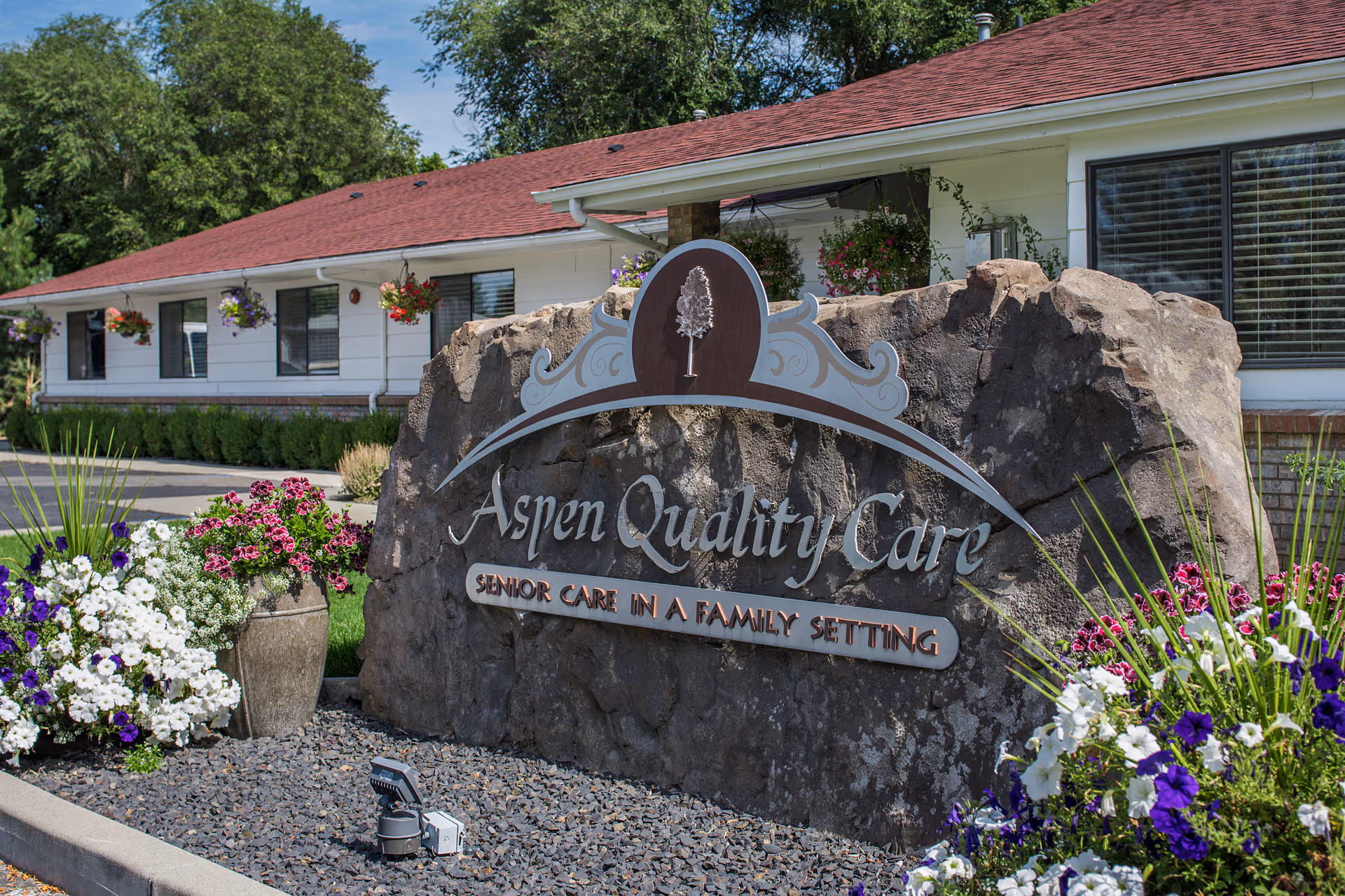A large stone sign in front of a single-story building with a red roof and white siding. The sign reads 'Aspen Quality Care Senior Care in a Family Setting' and is surrounded by colorful flowers and greenery. The building has several windows and hanging flower baskets.