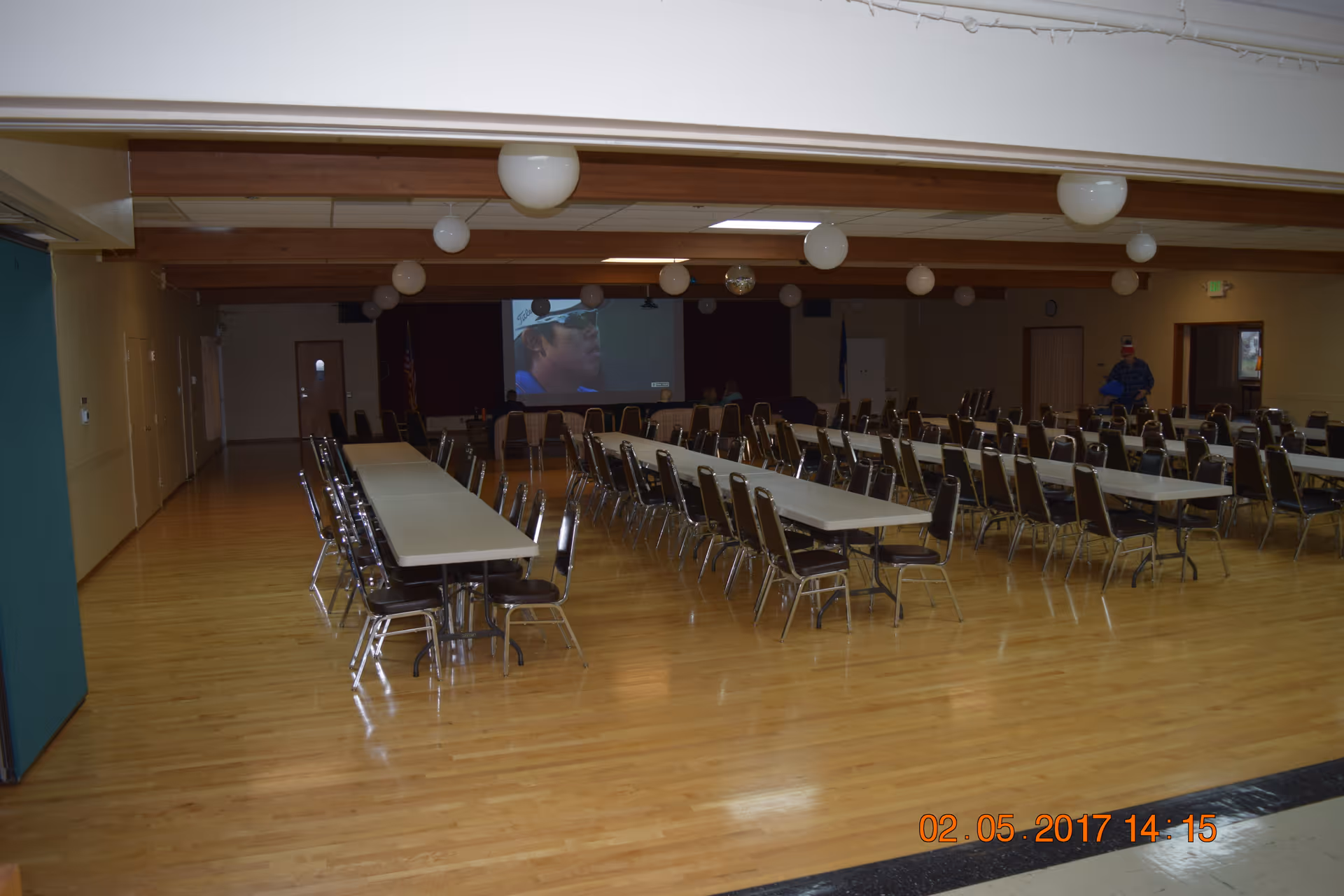 Large multipurpose dining/event hall with rows of long tables and many chairs facing a projection screen.