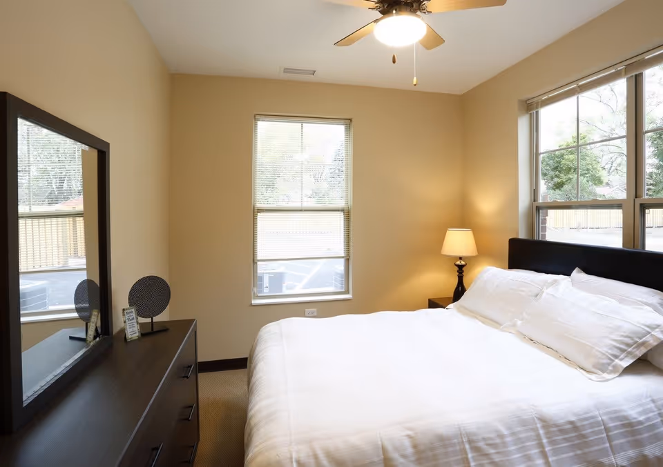 A bright bedroom with a bed made in white linens, a dresser with mirror, a bedside lamp, and two windows.