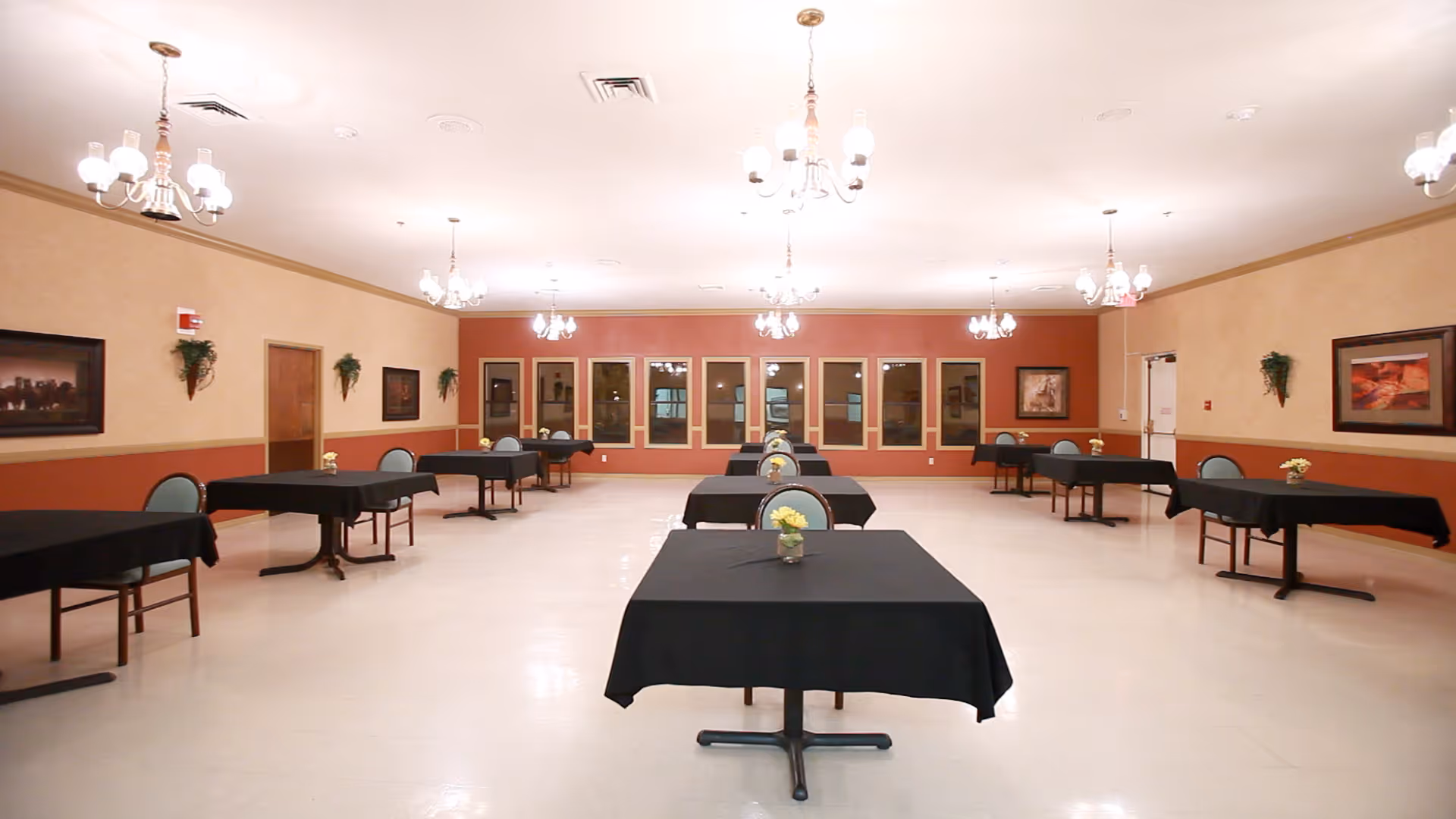 A large dining room with multiple tables covered in black tablecloths, each with a small flower arrangement. The room has beige and reddish walls, several framed pictures, and multiple chandeliers hanging from the ceiling providing warm lighting.