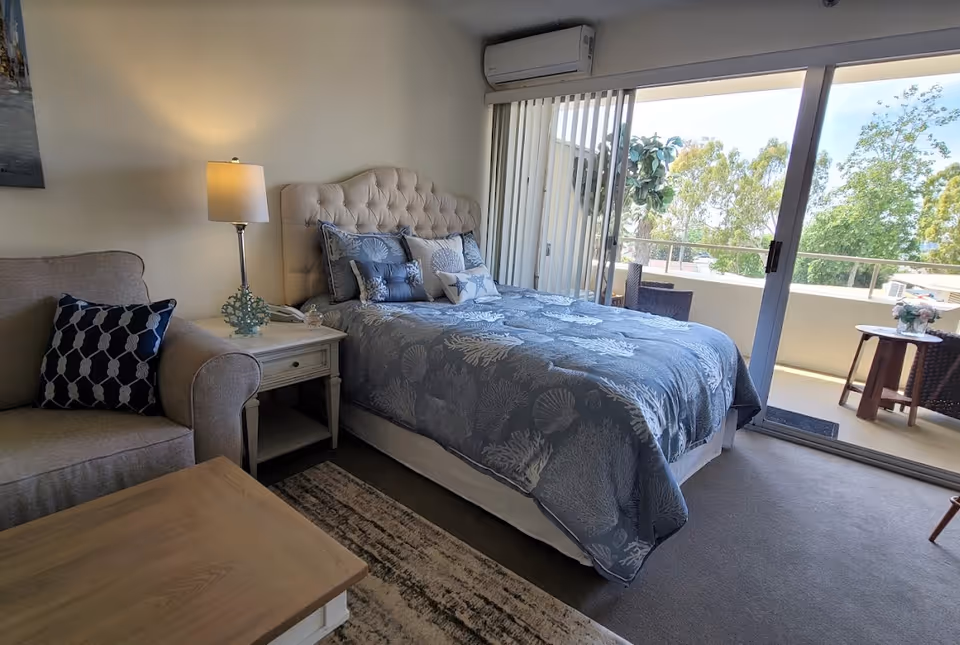 Well-lit bedroom with a tufted bed, nightstand and lamp, a small sofa, and sliding glass doors opening to a balcony.