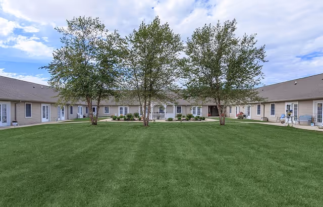 A large green lawn with three trees in the center, surrounded by a single-story building with multiple doors and windows, under a partly cloudy sky.