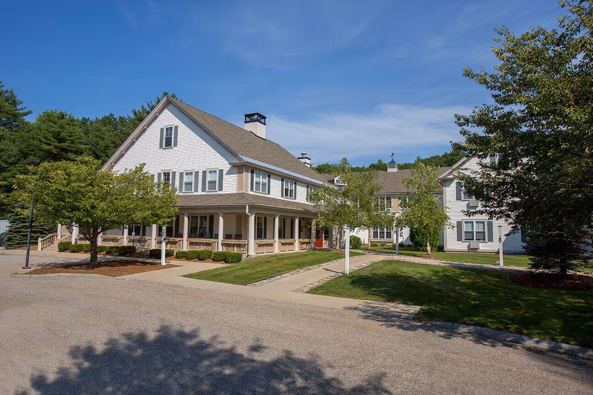 Exterior view of a large, white, two-story building with a pitched roof and multiple windows, surrounded by green trees and a well-maintained lawn under a clear blue sky.