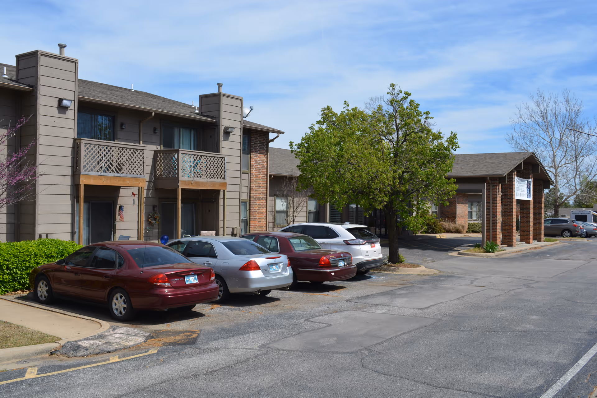 Exterior view of a residential building with parked cars in front, featuring balconies and a tree near the entrance under a partly cloudy sky.