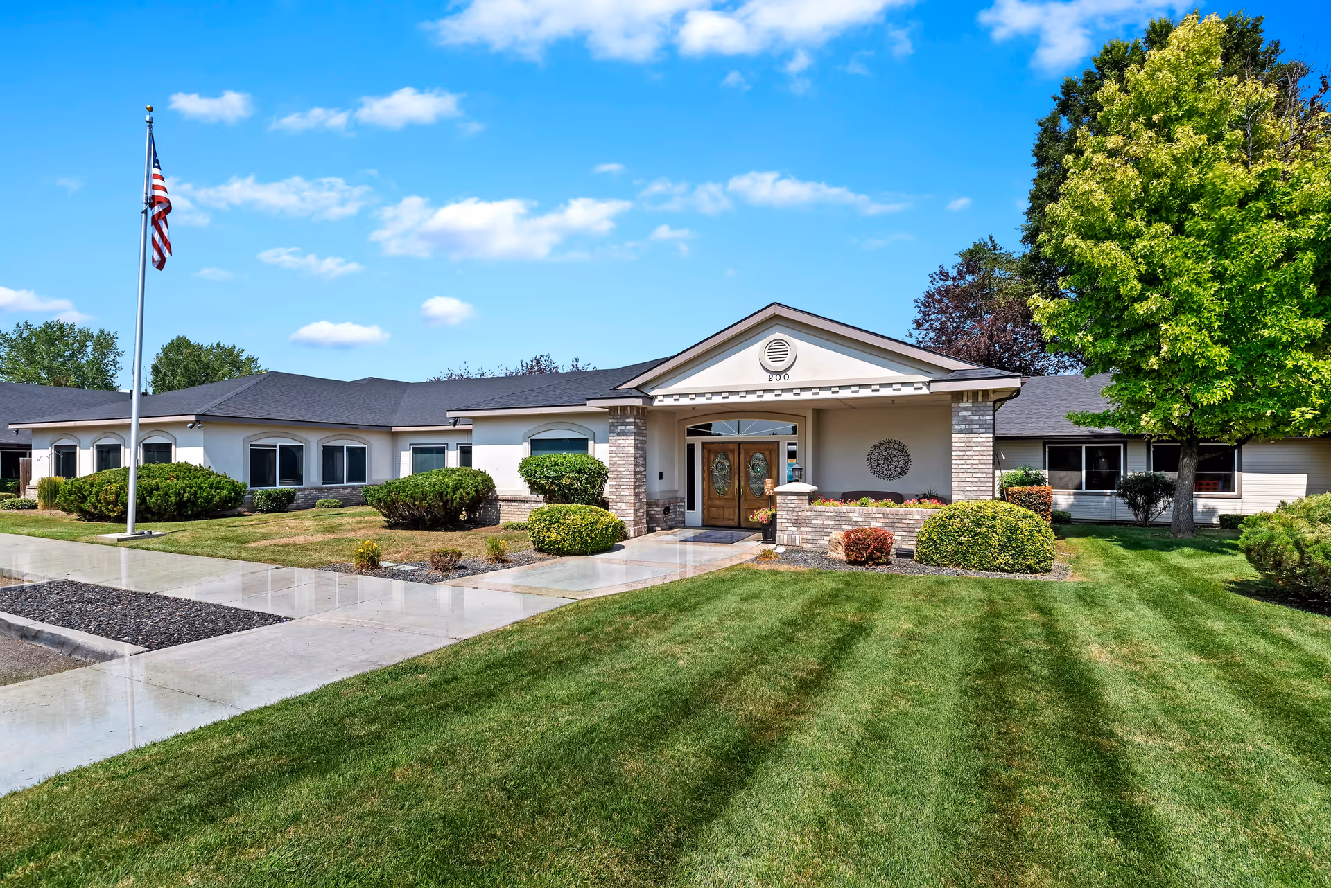 Front exterior view of a single-story assisted living facility building with a well-maintained lawn, shrubs, a large tree, and an American flag on a flagpole under a blue sky with scattered clouds.