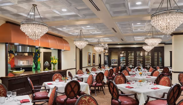 Elegant dining room with multiple round tables covered with white tablecloths, each set with glassware, plates, and red napkins. The chairs have red and gold patterned upholstery. Crystal chandeliers hang from a coffered ceiling, and a wood-fired oven is visible in the background behind a counter with decorative items and flowers.