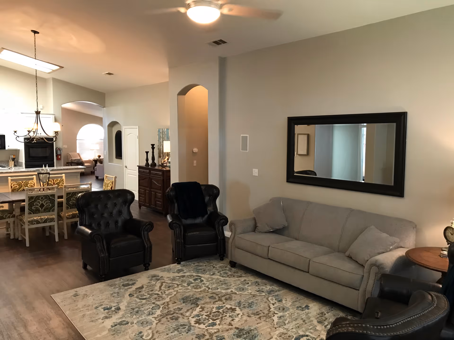 Open-plan living room with a gray sofa, two black leather armchairs, a patterned rug, and a dining area and kitchen visible in the background.