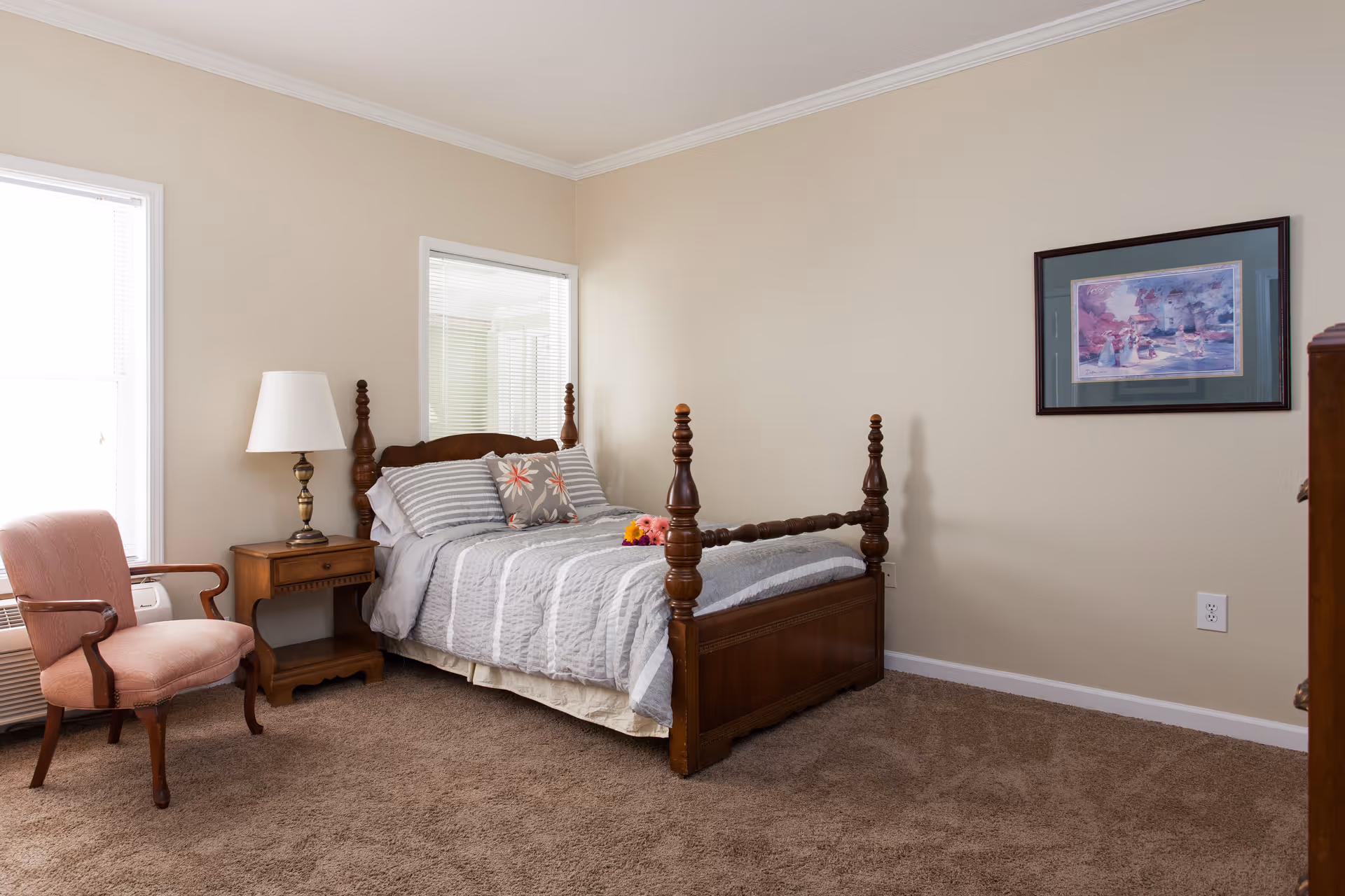 A cozy bedroom with a wooden four-poster bed made with gray and white striped bedding and decorative pillows. Next to the bed is a wooden nightstand with a brass lamp. A pink upholstered armchair is placed near a window with blinds. The room has beige walls, carpeted floor, and a framed painting on the wall.