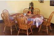 A dining area with a rectangular wooden table covered with a checkered tablecloth and surrounded by six wooden chairs. There is a small decorative centerpiece on the table and a wall-mounted shelf with decorative items in the background.