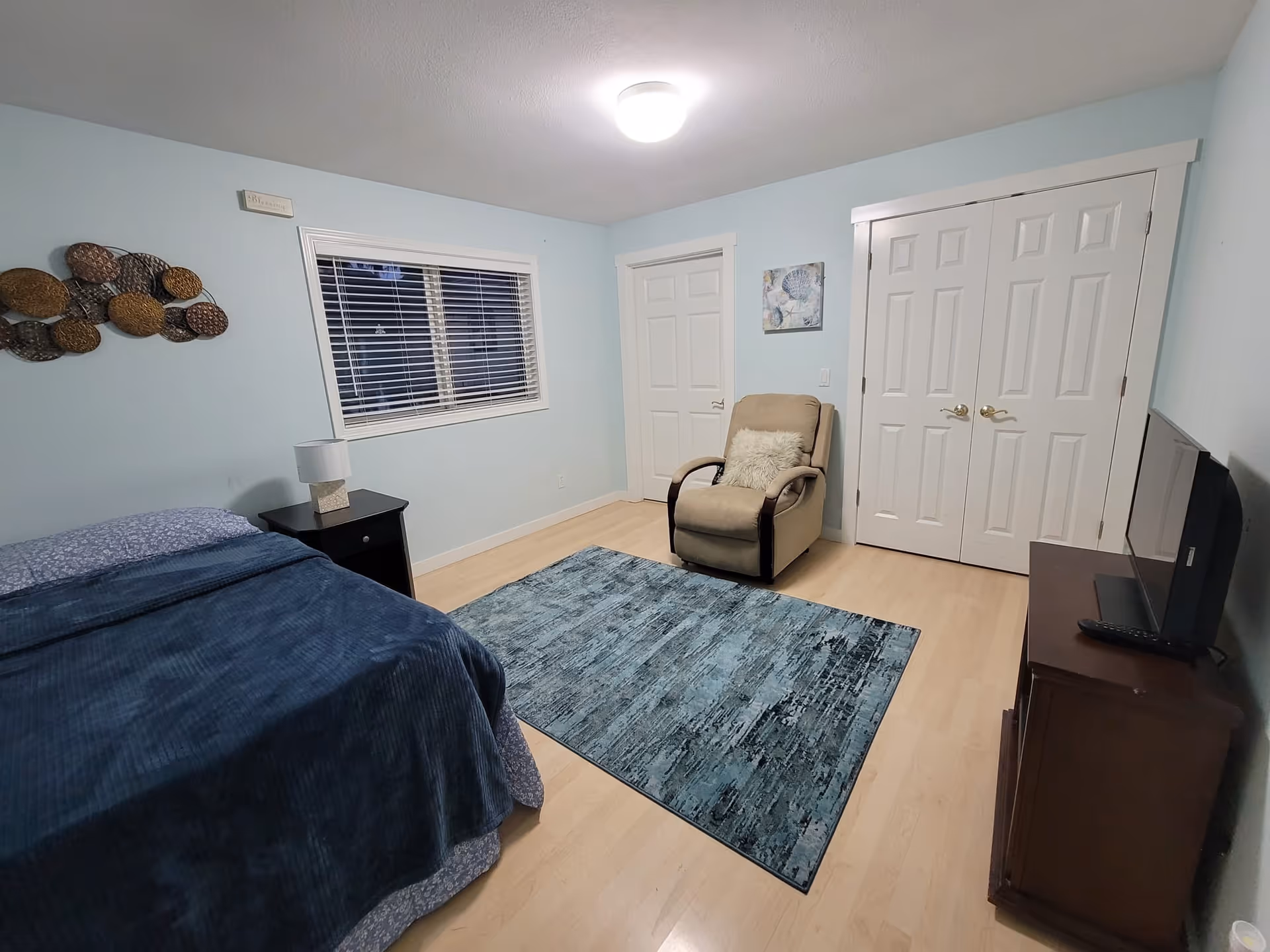 A bedroom with light blue walls and light wood flooring. The room contains a bed with a blue blanket, a black nightstand with a lamp, a beige armchair with a white cushion, a blue patterned rug, a flat-screen TV on a wooden stand, and two white doors including a closet with double doors. There is a window with white blinds and decorative wall art.