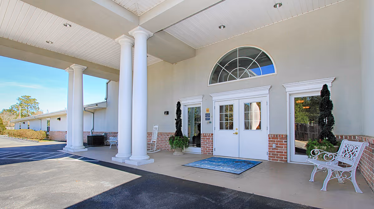 Entrance of Gulf Breeze Courtyard Assisted Living featuring white double doors with glass panels, flanked by tall white columns and decorative plants, with a white bench on the right side and a covered driveway area.