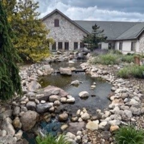 A landscaped rock-lined pond and stream leading toward a stone-faced building with trees and a cloudy sky.