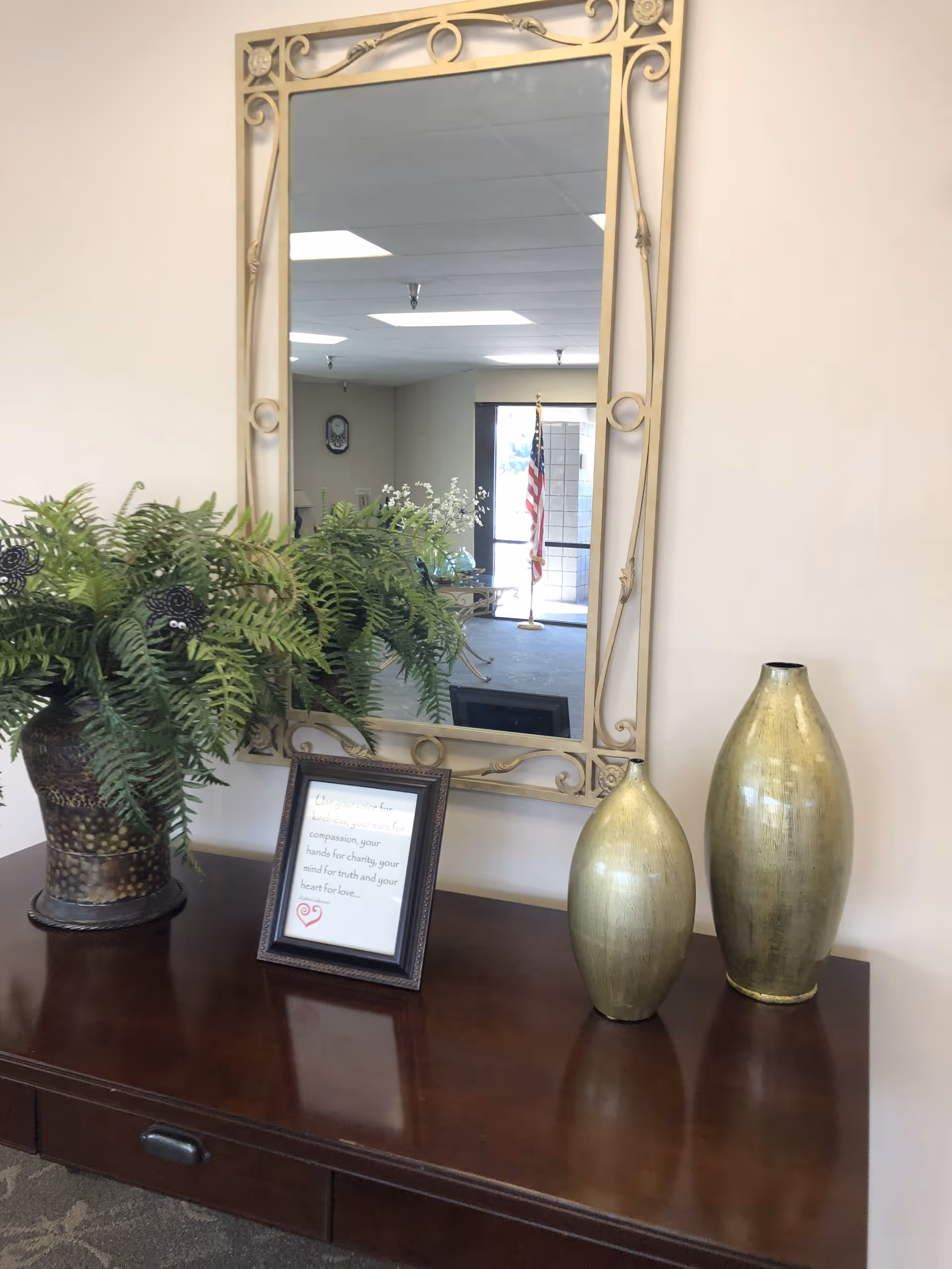 Wood console table topped with a potted fern, framed sign and two gold vases beneath a large ornate mirror reflecting an American flag and lobby entrance.