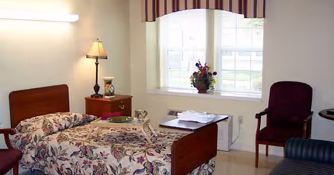 A single bed with floral bedding in a well-lit room. Next to the bed is a wooden nightstand with a lamp and a small decorative item. A window with blinds and a valance lets in natural light, and a vase with flowers is placed on the windowsill. There is a maroon upholstered chair and part of a striped sofa visible in the room.